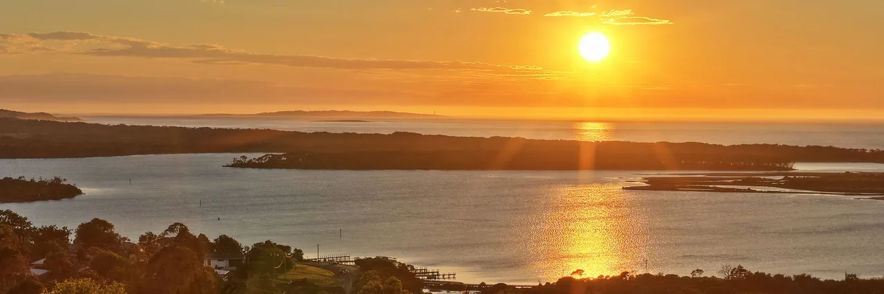 Aerial panorama of calm Mallacoota Lake in warm golden sunrise light at 6:16 am AEDT on 25 October 2026.